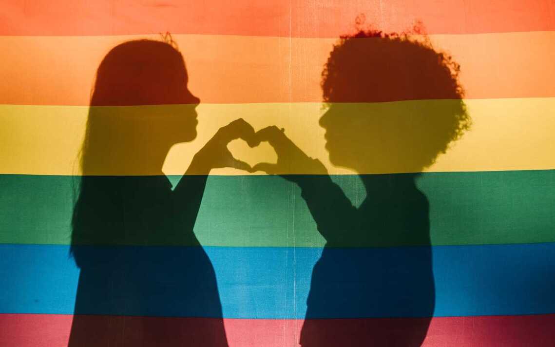 Silhouette of two women making a heart sign together behind a rainbow flag 