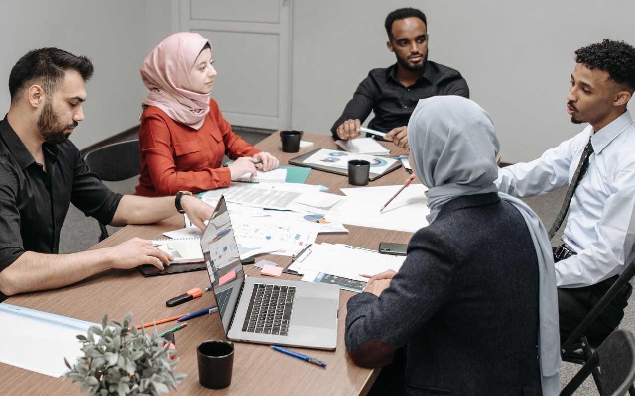 Group of diverse people sitting and talking  around a table with papers, pens and laptops