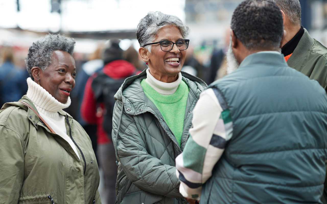 A black woman with short grey hair listens as a medium-skinned older woman with short grey hair smiles and greets two men with people blurred in the background at an event